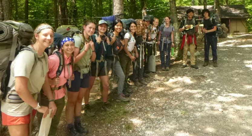 A group of students wearing backpacks stand in a line and smile for the photo. 